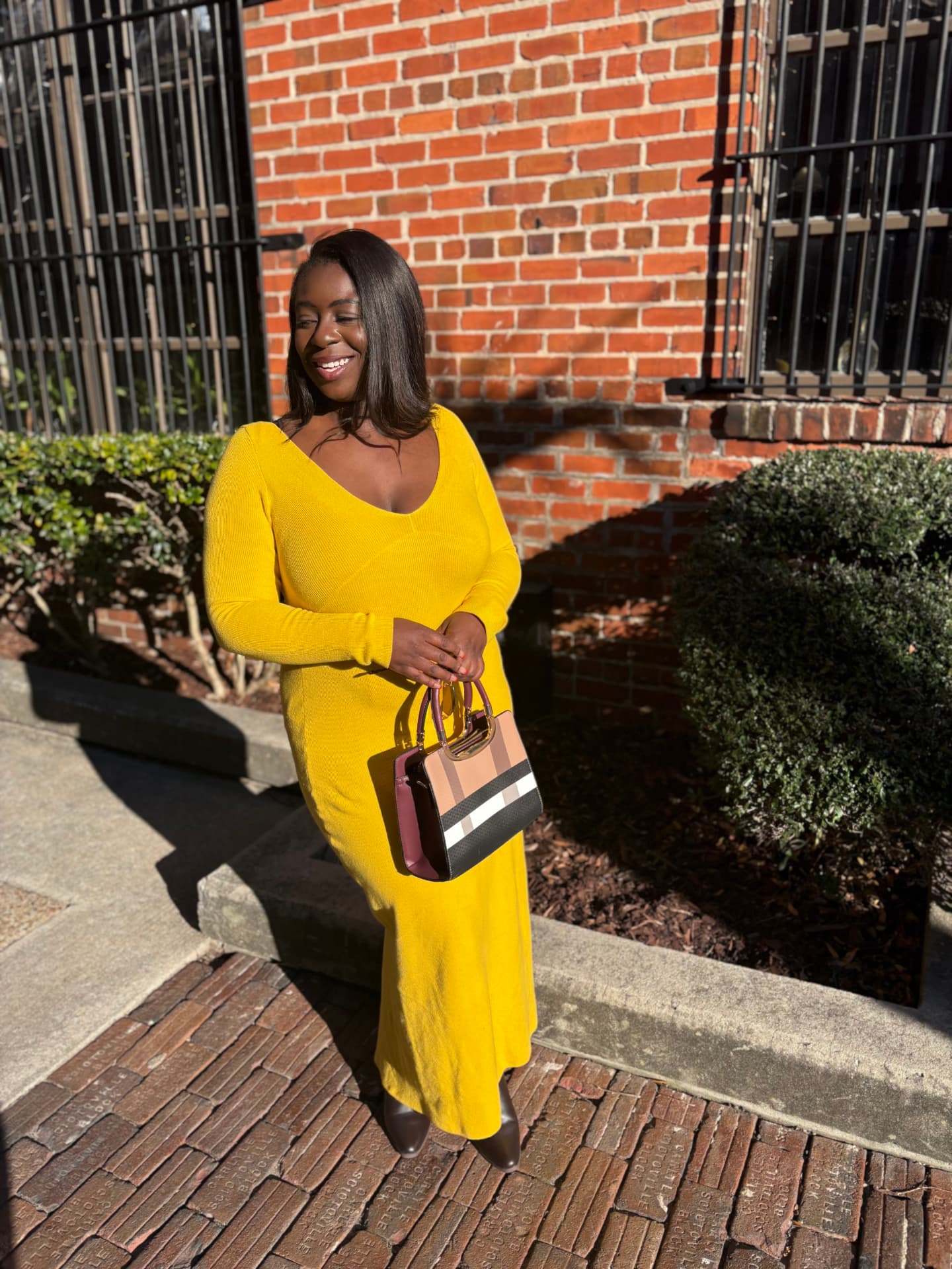 Moriah Davis in a long yellow knit dress holding a plaid handbag on a brick sidewalk