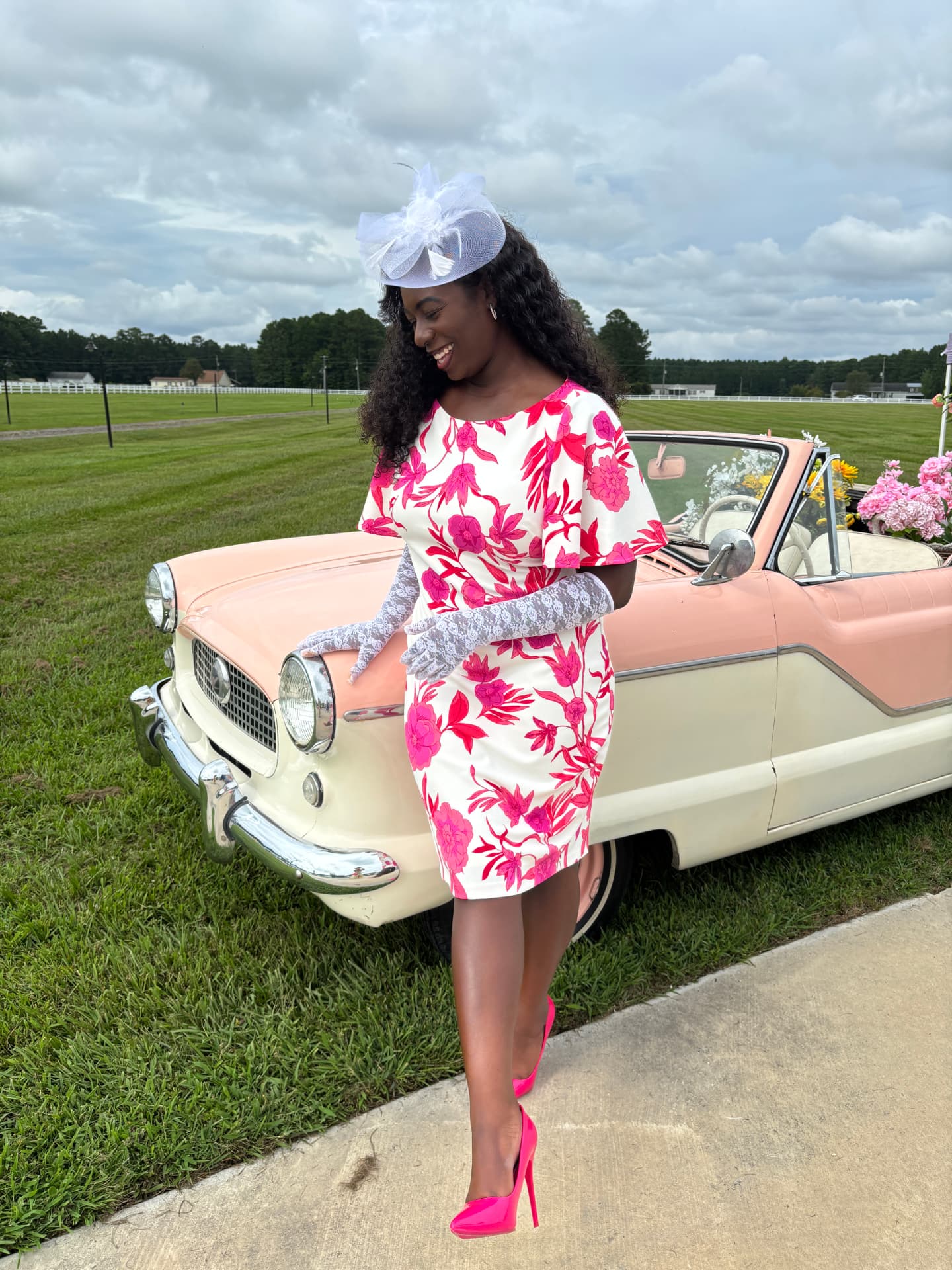Moriah Davis in a pink floral dress and white fascinator beside a vintage pink convertible